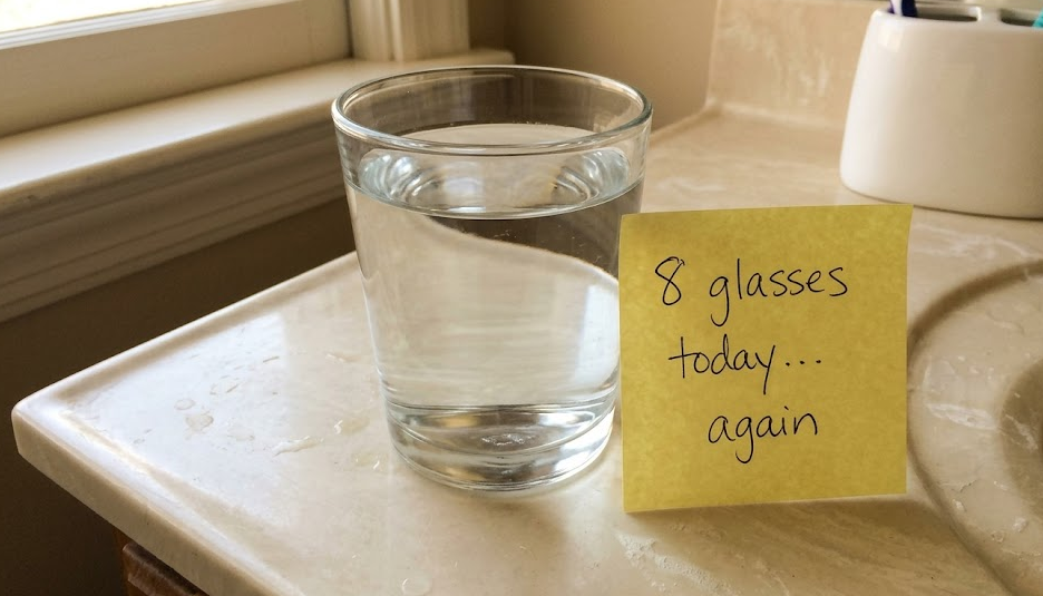 A glass of water on a bathroom counter, morning light, with a sticky note reading 8 glasses today again